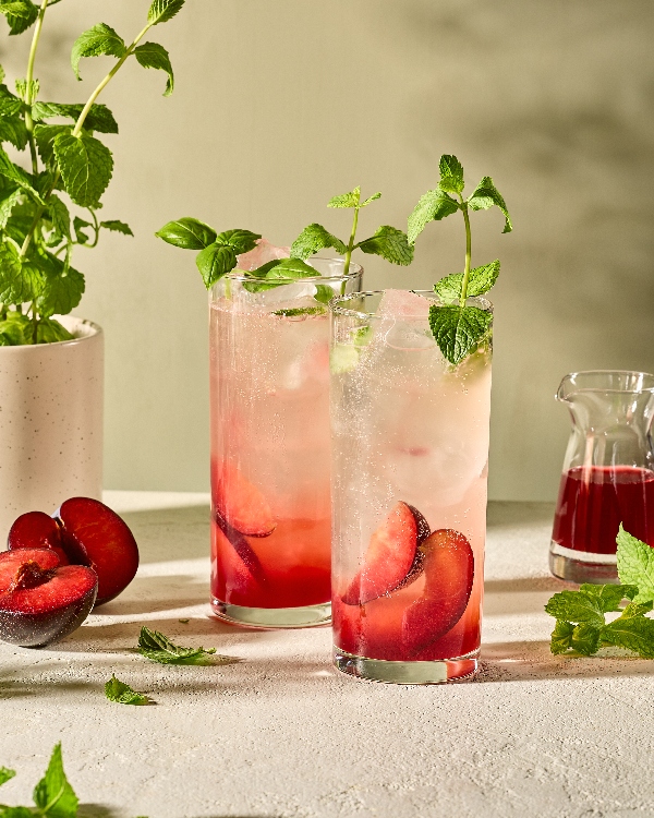 Two tall glasses filled with a plum and herb-infused sparkling drink, garnished with fresh mint and basil. The beverages are layered with vibrant red plum slices at the bottom and pale, fizzy liquid above. Surrounding the glasses are fresh mint sprigs, halved plums, a white planter with mint, and a small glass pitcher of deep red syrup, all set on a light, textured surface with soft natural lighting.