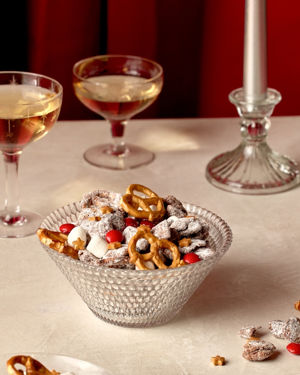 A clear glass bowl filled with a mix of pretzels, marshmallows, chocolate candies, and powdered sugar sits on a light-colored surface. In the background, two glasses of what appears to be champagne and a glass candle holder with a white candle are visible.