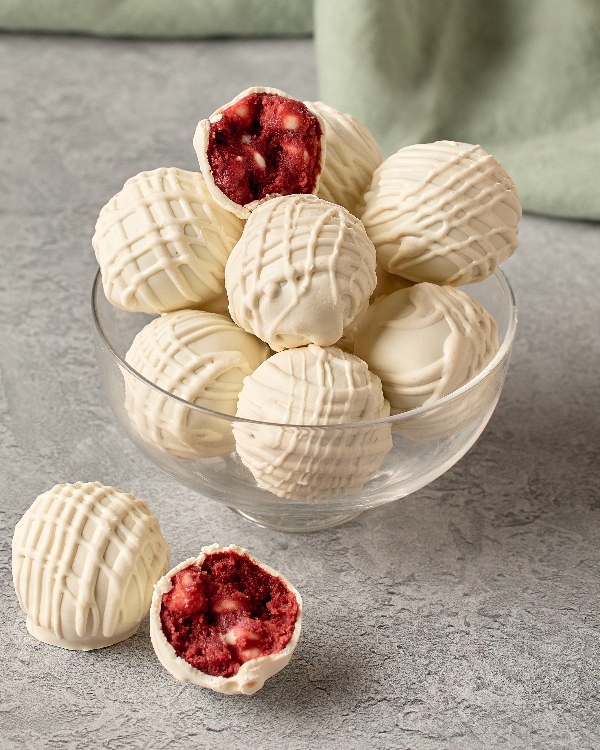 A glass bowl of red velvet cookie dough truffles dipped in white chocolate with drizzle. One is shown cut in half, with the red centre visible.