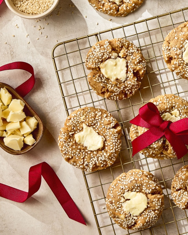 White chocolate chunk tahini cookies coated in sesame seeds cooling on a wire rack, with one cookie tied in a red ribbon. A bowl of sesame seeds, a dish of chopped white chocolate, and loose red ribbon are arranged around the cookies on a light textured surface.