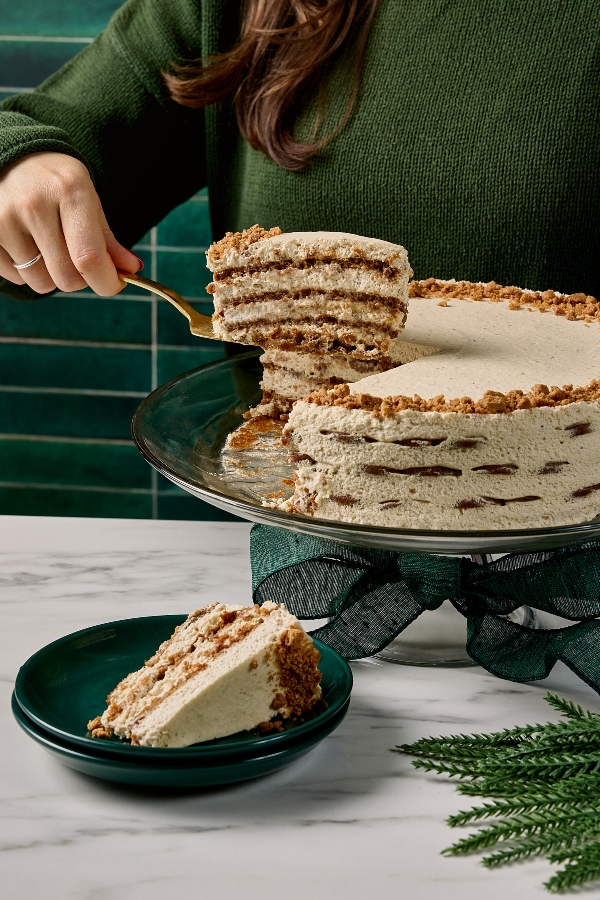 A person in a green sweater lifts a tall slice of gingersnap icebox cake from a cake stand using a gold server, revealing its stacked layers of spiced cookie crumbs and creamy filling. A plated slice sits on a dark green dish below, with evergreen sprigs on the marble surface.