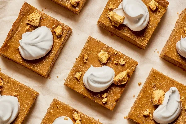 Overhead view of neatly arranged pumpkin dessert bars on parchment paper. Each square is topped with a swirl of whipped cream and a sprinkling of golden, crunchy crumble pieces. The bars have a rich, dark orange hue.