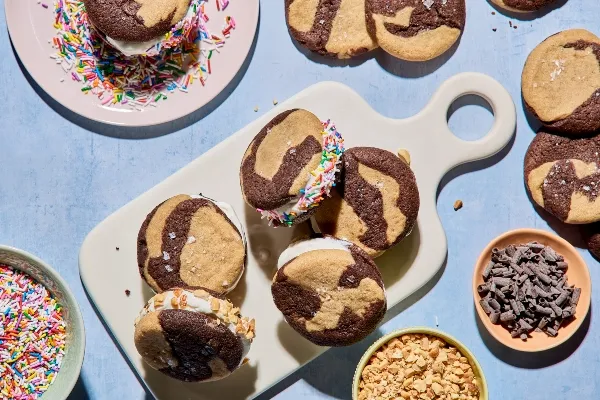 A platter of marbled chocolate and peanut butter ice cream sandwiches on a white serving board, some rolled in rainbow sprinkles or chopped nuts. More cookies and bowls of toppings surround the scene on a light blue surface.