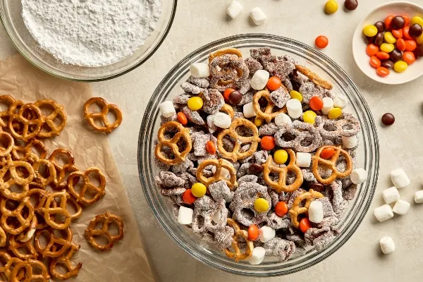 Overhead view of a large glass bowl filled with Puppy Chow, a chocolate-coated cereal and pretzel snack dusted in icing sugar. Surrounding the bowl are scattered pretzels on parchment paper, a bowl of icing sugar, and a small dish of chocolate chips.
