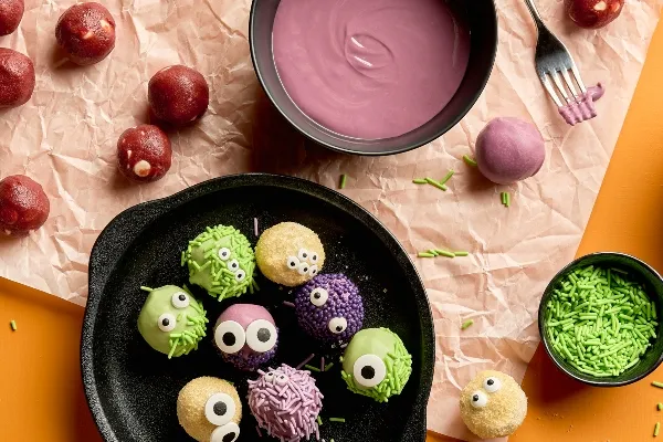 Halloween-themed red velvet cookie dough truffles decorated as monster faces, coated in green, purple, or pink candy melts, then rolled in sprinkles or sugar and topped with candy eyes. A black dish holds the finished truffles, and uncoated red velvet balls, a bowl of purple coating, green sprinkles, and a fork rest on crumpled parchment nearby on a bright orange surface.