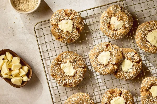 A cooling rack filled with freshly baked tahini cookies coated in sesame seeds, each topped with melted white chocolate. A small bowl of sesame seeds and a dish of chopped white chocolate sit beside the rack on a light stone surface.