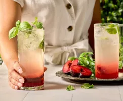 A person in a cream-colored linen outfit holding a tall glass of a layered plum cocktail garnished with fresh basil and mint. The drink features a deep red fruit layer at the bottom and a pale sparkling top. On the table are more glasses, fresh herbs, and halved plums on a silver tray, all set against a lush green backdrop