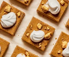 Overhead view of neatly arranged pumpkin dessert bars on parchment paper. Each square is topped with a swirl of whipped cream and a sprinkling of golden, crunchy crumble pieces. The bars have a rich, dark orange hue.