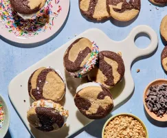 A platter of marbled chocolate and vanilla ice cream sandwiches on a white serving board, some rolled in rainbow sprinkles or chopped nuts. More cookies and bowls of toppings surround the scene on a light blue surface.