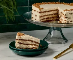 A layered gingersnap icebox cake on a glass cake stand, with visible layers of spiced cookie crumbs and creamy filling. A slice is served on a dark green plate in the foreground, alongside a gold cake server on a marble countertop.