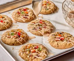 Freshly baked fruitcake cookies on a parchment-lined baking sheet, topped with colourful candied fruit and being drizzled with a glossy glaze from a spoon. A bowl of glaze sits nearby.