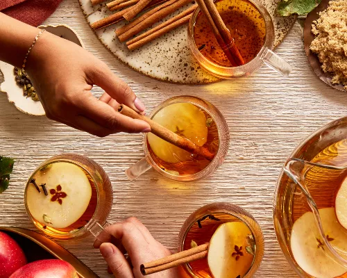 Four glass mugs of hot apple cider garnished with apple slices, cloves, and cinnamon sticks, shown on a table with a pitcher of cider, a bowl of apples, and a bowl of golden yellow sugar, and hands reaching for the mugs.