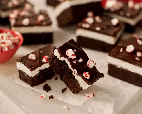 A close-up of chocolate mint brownies with a layer of mint cream and ganache top, each sprinkled with candy cane pieces, shown on wax paper on a white surface. 