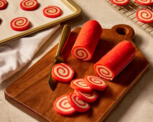  Two rolls of red and white peppermint pinwheel cookie dough on a wooden cutting board with several slices cut, with more slices on a baking sheet and baked cookies cooling on a wire rack.