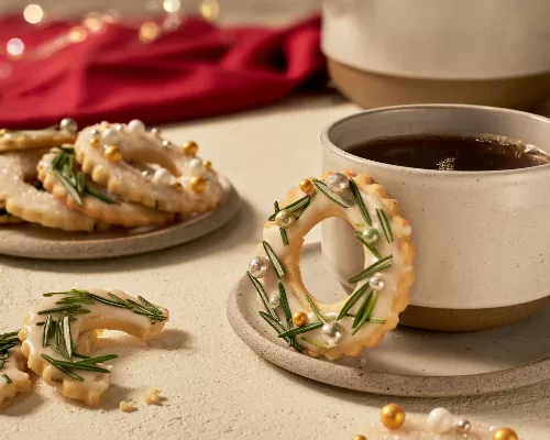 Glazed sugar cookies with icing and decorated like wreaths with rosemary, one on a saucer with a cup of coffee, one broken on a table, and several on a plate, shown in a festive holiday setting.