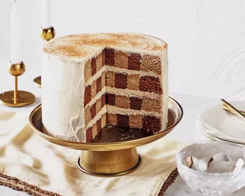 Eggnog and spiced cocoa checkered cake with slices missing on a brass cake stand, shown with candles, plates, and gold utensils