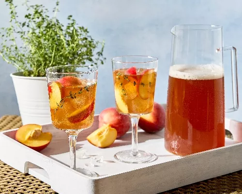 A white tray on a wicker table holds two stemmed glasses, a pitcher of peach-thyme sweet tea, and whole and cut fresh peaches. The drinks are garnished with fresh peach slices and sprigs of thyme, and a potted thyme plant is visible in the background.